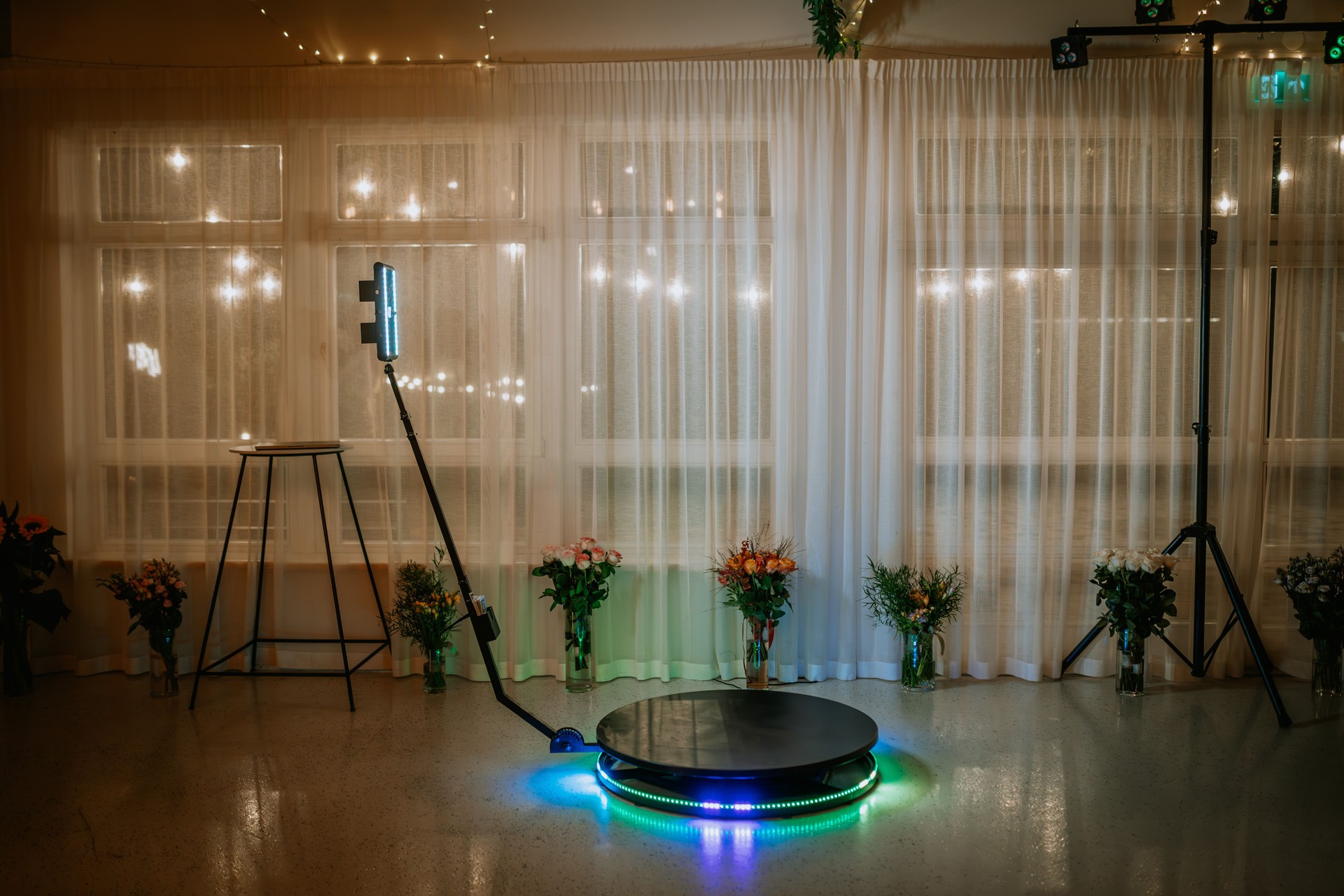 Une configuration de photomaton à 360 degrés avec des lumières LED et une plate-forme rotative, entourée d’arrangements floraux, devant des rideaux transparents et des guirlandes lumineuses.